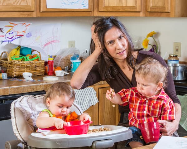 Mom with a toddler and baby in a high chair in a messy kitchen. Mom has her hand on her head and her hair is disheveled.