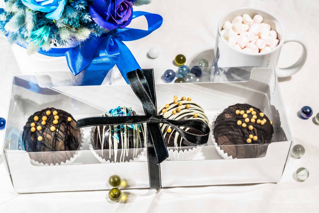 Four hot chocolate bombs in a gift box with a blue bow and a mug with marshmallows above it.