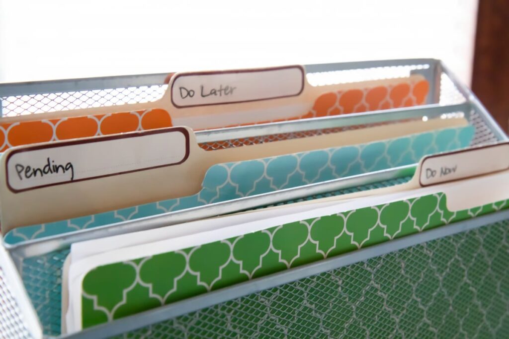 A metal file holder with three folders in it, one called Do Now, one Pending, and one Do Later for a paper organization system.