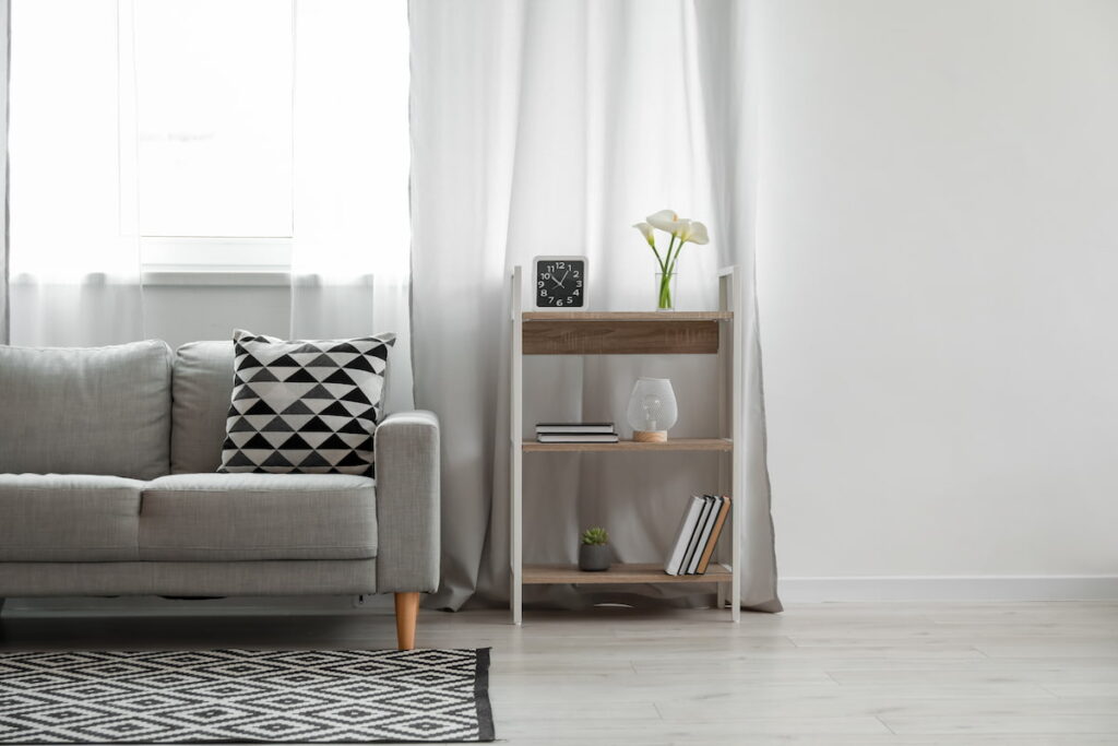 Tidy neutral colored living room showing the edge of a sofa with a small side shelf next to it and a window in the background.