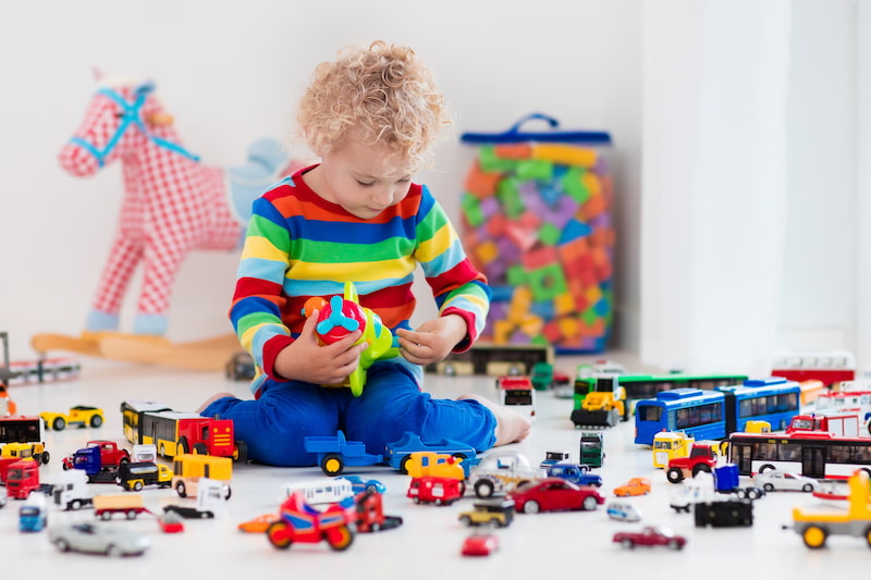 Little boy playing with toys on the playroom floor. Mostly little cars and trucks with a rocking horse in the background.