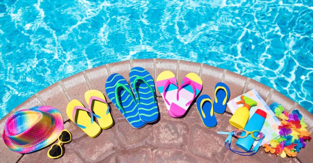 The curved corner of a pool, with water above and poll accessories sitting on the edge, including four colorful pairs of flip flops.