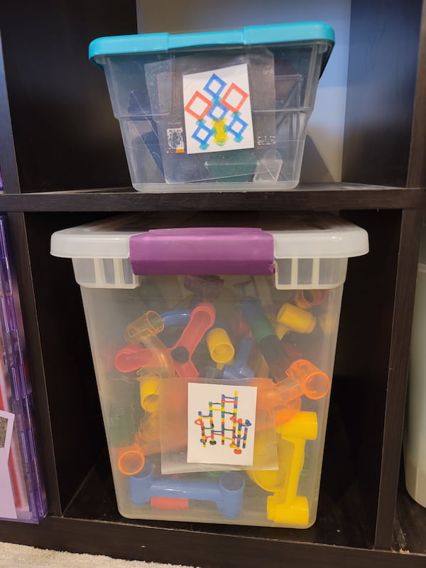 Toy shelf with two clear bins for toys with picture labels on them. One bin has a picture of stacking blocks and the other bin has a picture of a marble run.
