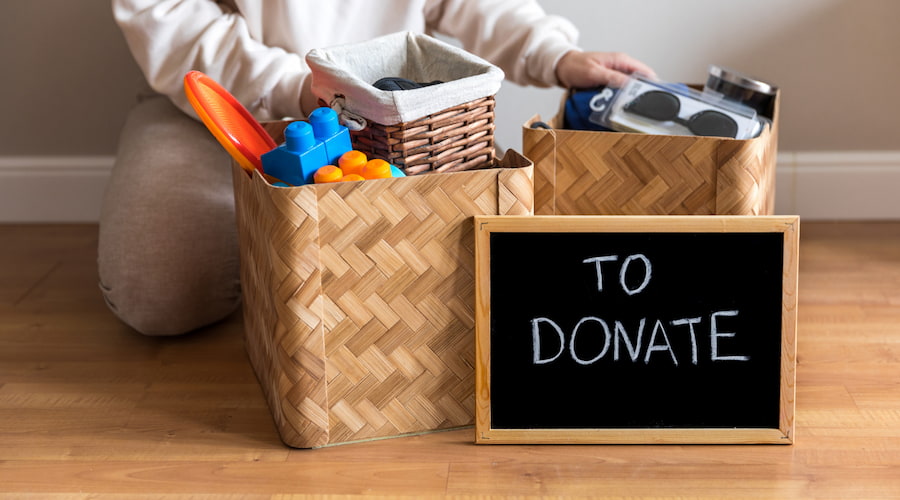 Neck down body kneeling behind a basket of clutter with a chalkboard in front that says,"to donate."