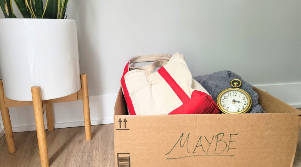 A Maybe Box used for decluttering things one is not sure if they want to get rid of. This box is labeled maybe and has a bag, a blanket, and a clock in it, with a plant in the background.