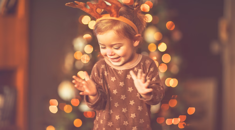 Little girl with antlers standing in front of a lit up Christmas tree.